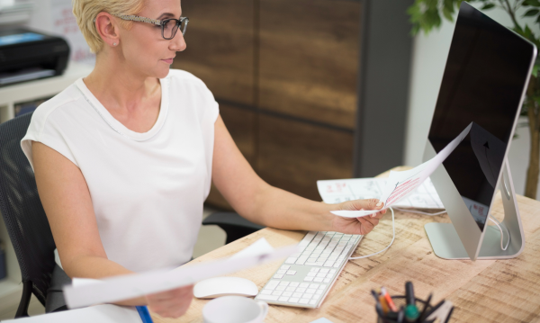 Woman reviewing paperwork at a desk while working on a computer.
